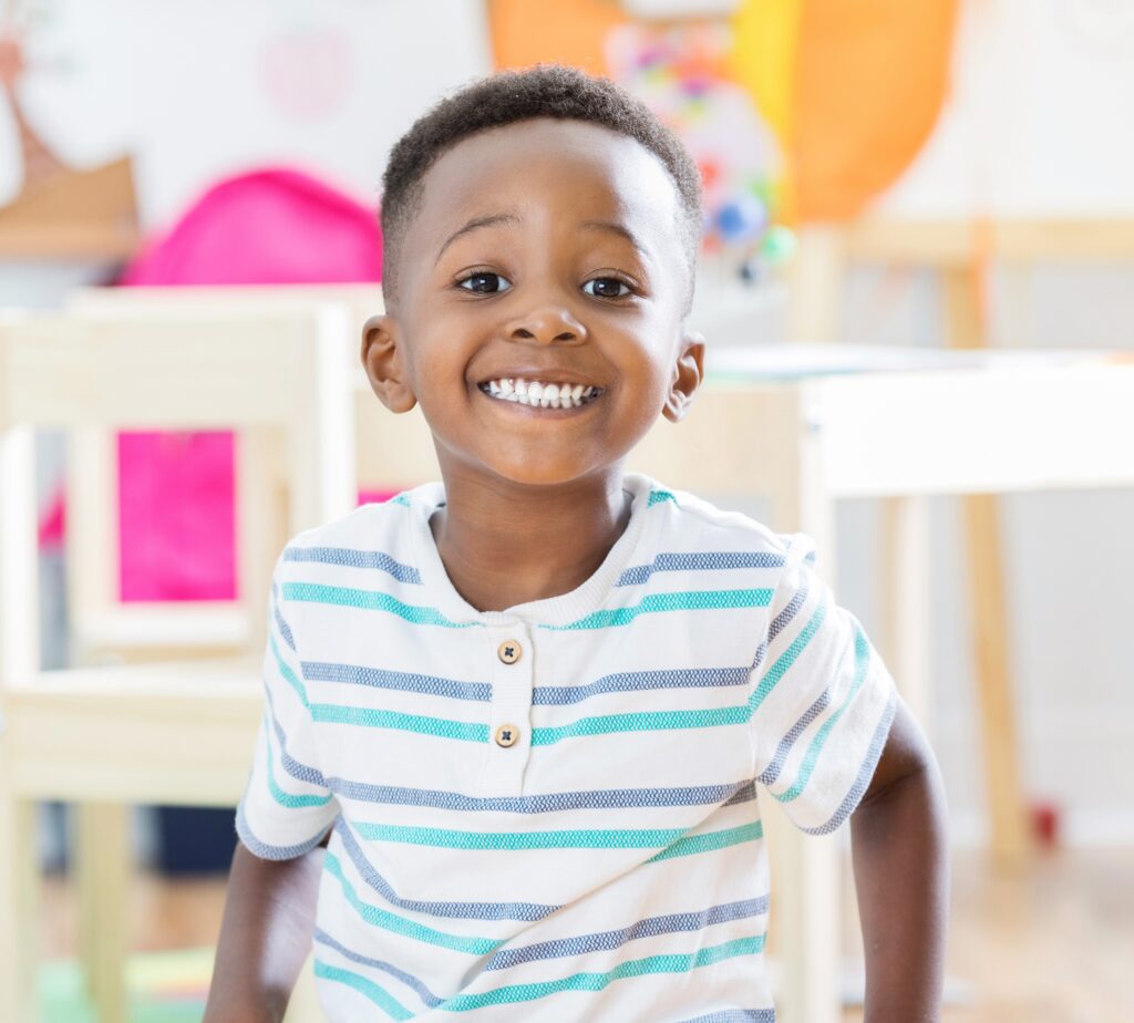 Adorable Preschool Age Little Boy Sits On Floor Of His Classroom And Smiles For Camera